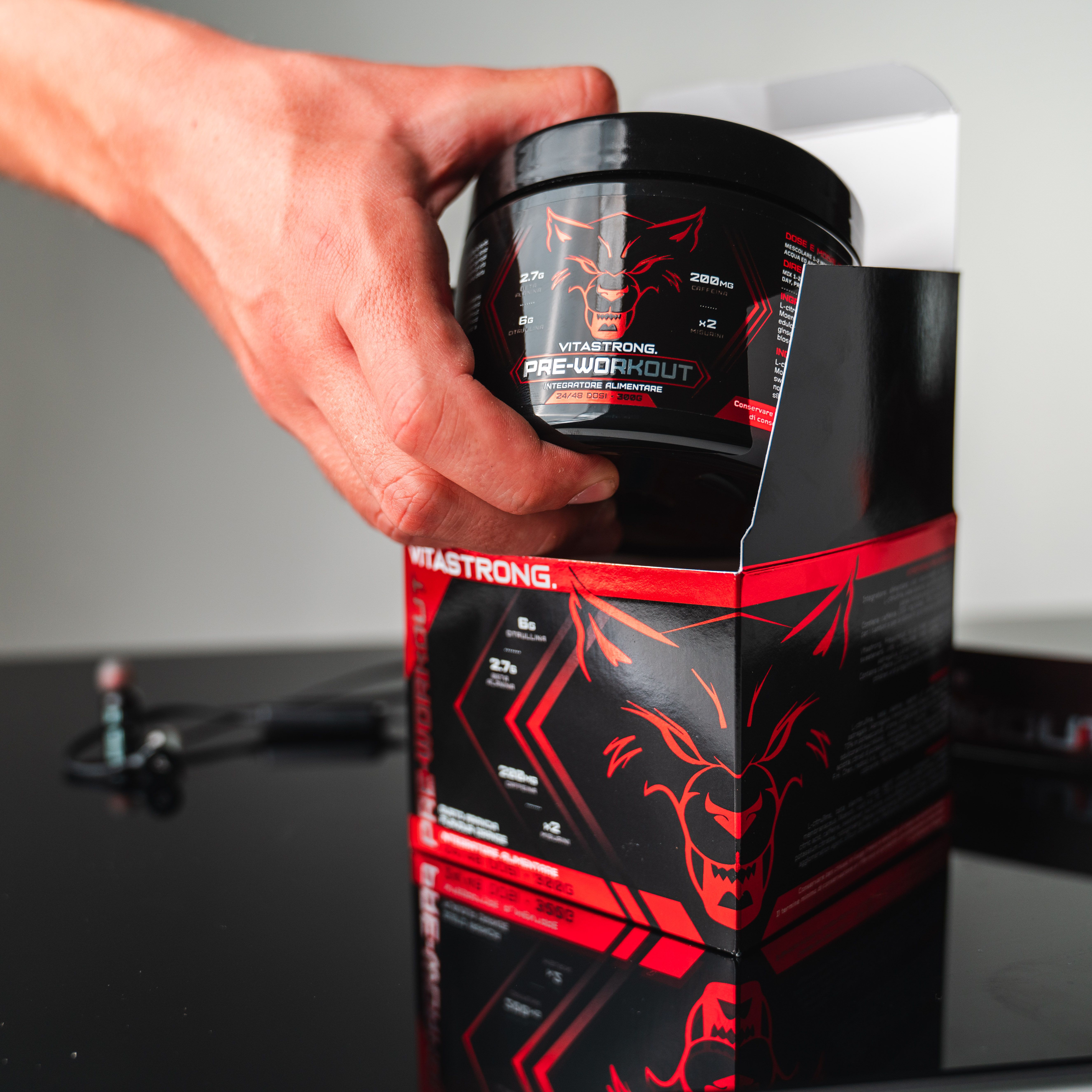Main ouvrant une boîte Vitastrong Pre-Workout. Pot noir avec logo rouge. Fond noir.