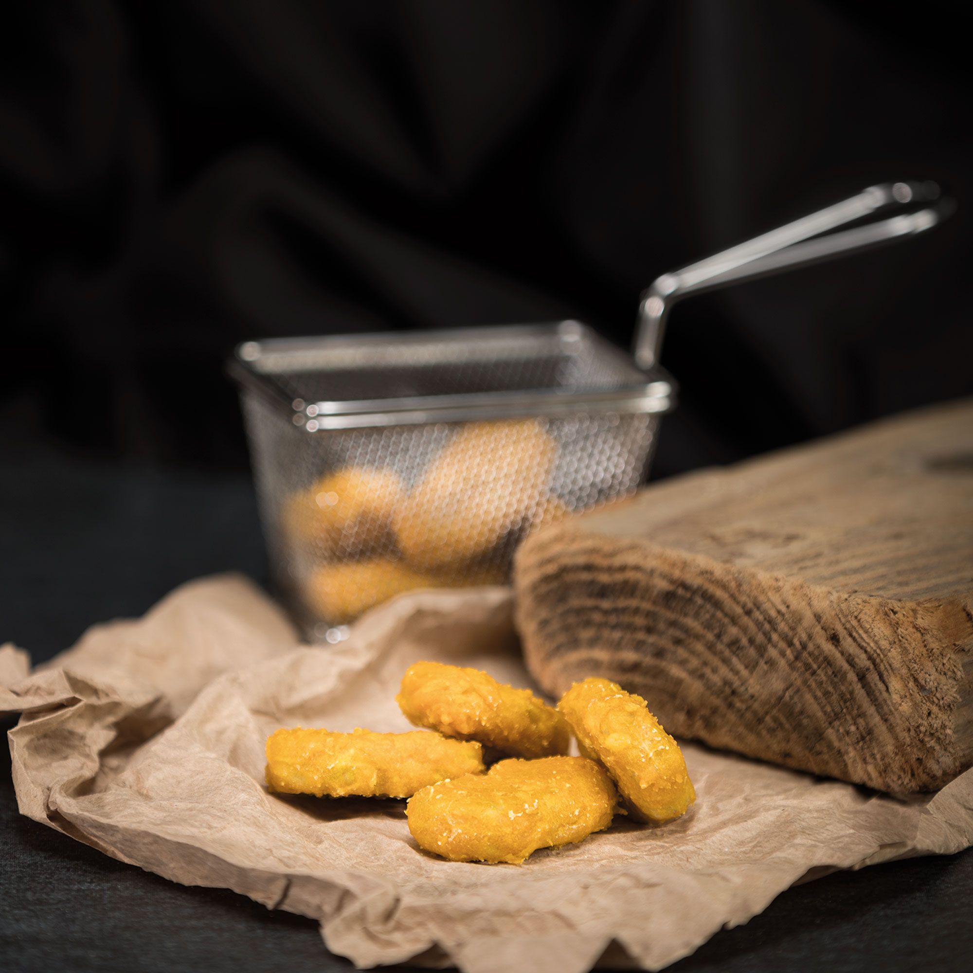 Chicken Nuggets in einem Metallkorb auf Papier. Einige Nuggets liegen daneben. Holzbrett im Hintergrund.