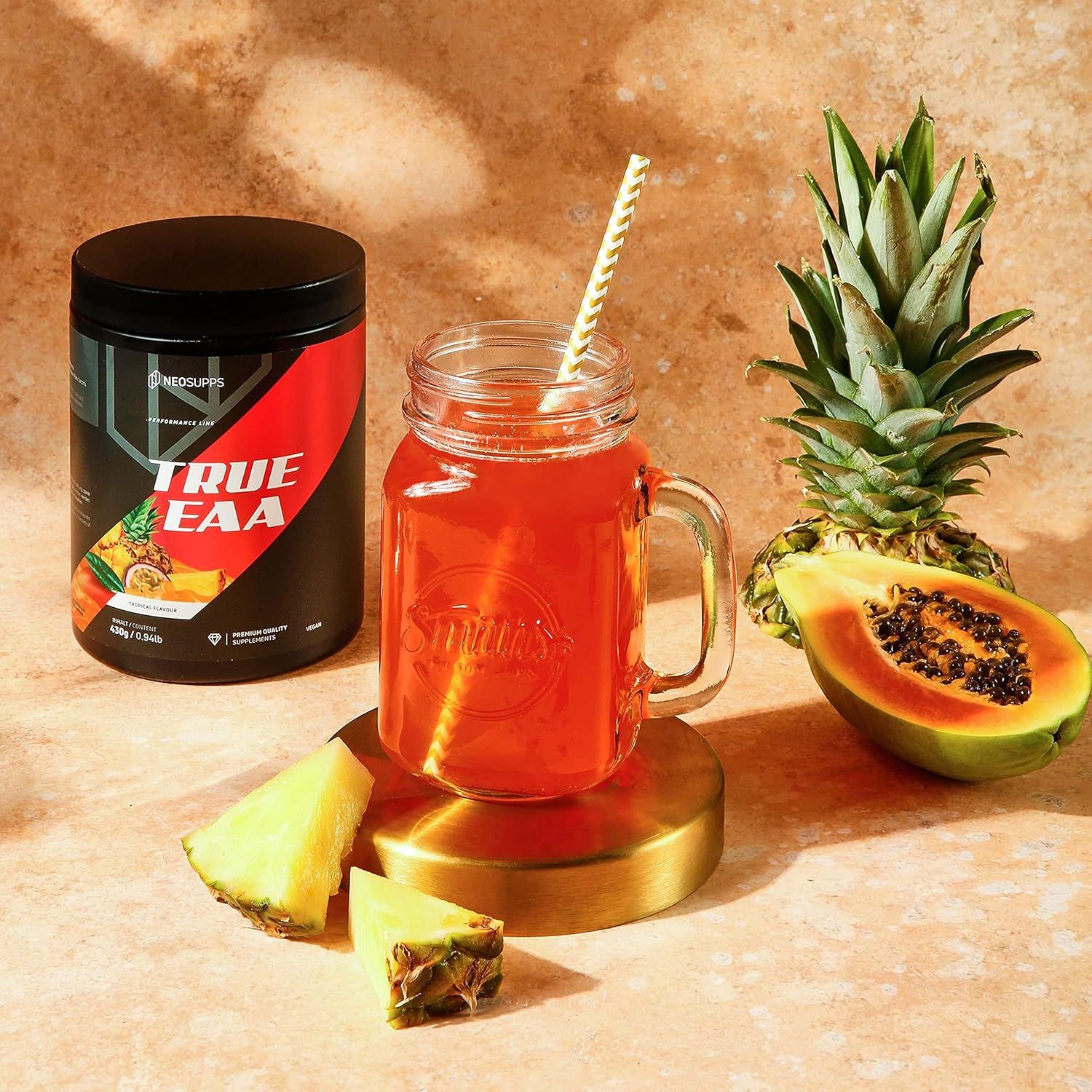 Schwarze Dose TRUE EAA neben Glas mit Getränk, Ananas, Papaya und Ananasstücken. Goldene Untersetzer.