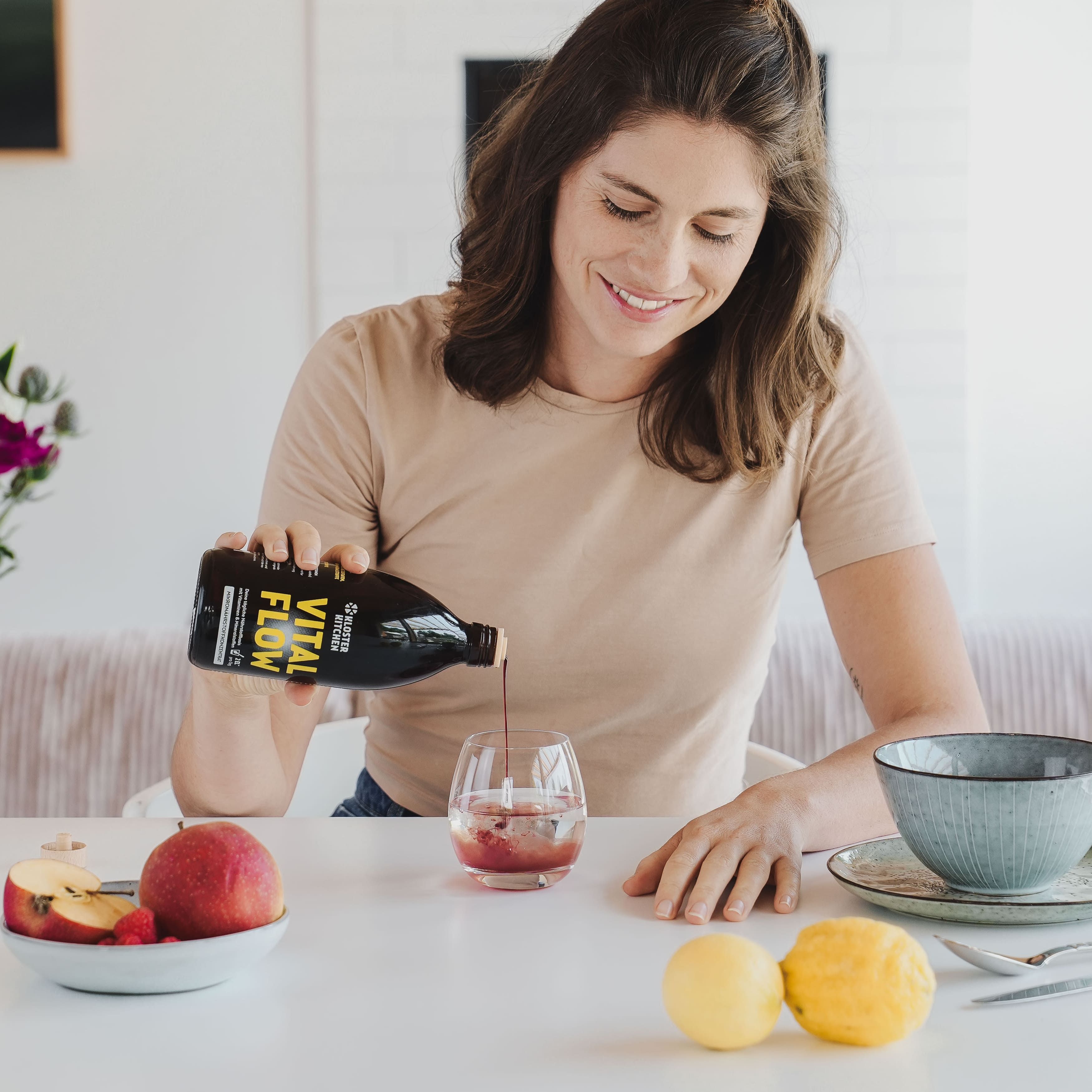 Frau gießt Flüssigkeit aus Flasche VITAL FLOW in ein Glas. Auf Tisch: Obst, Schüssel.