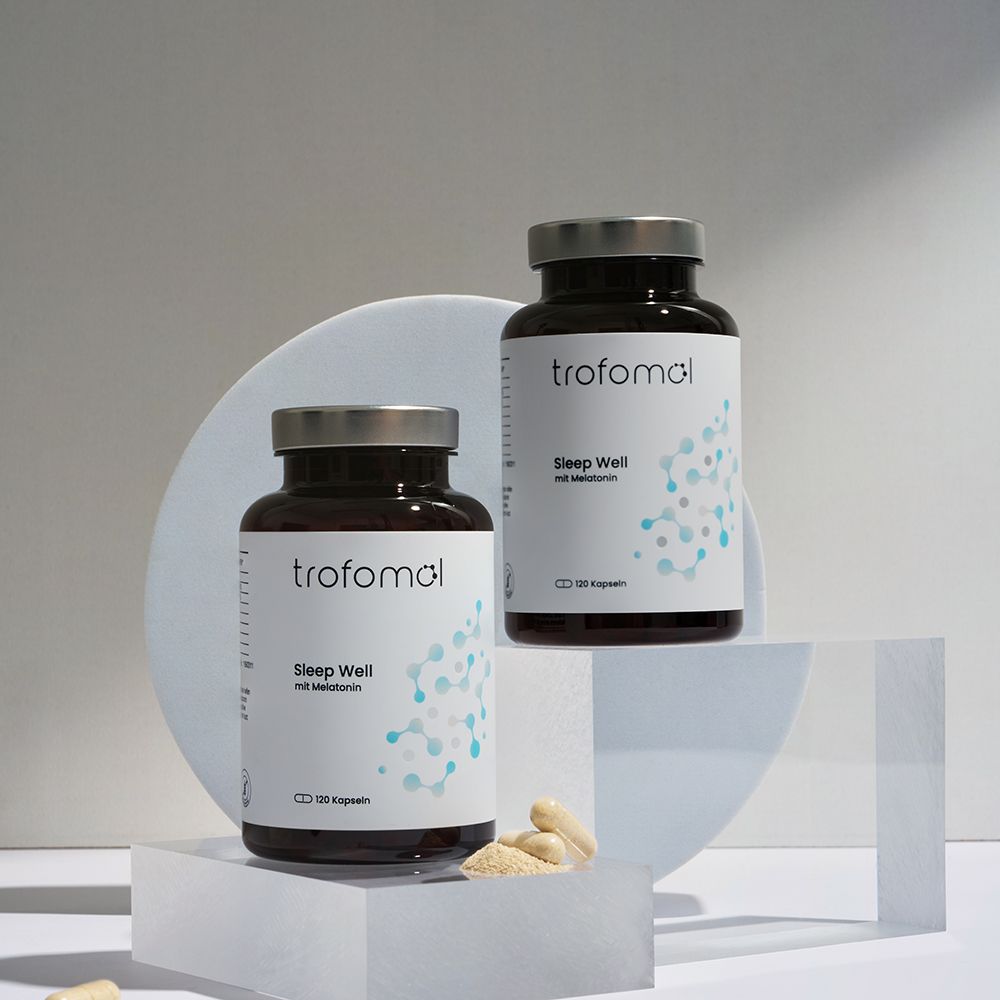 Zwei braune Glasflaschen mit silbernen Deckeln. Weiße Etiketten mit "trofomol Sleep Well mit Melatonin". Einige Kapseln daneben.