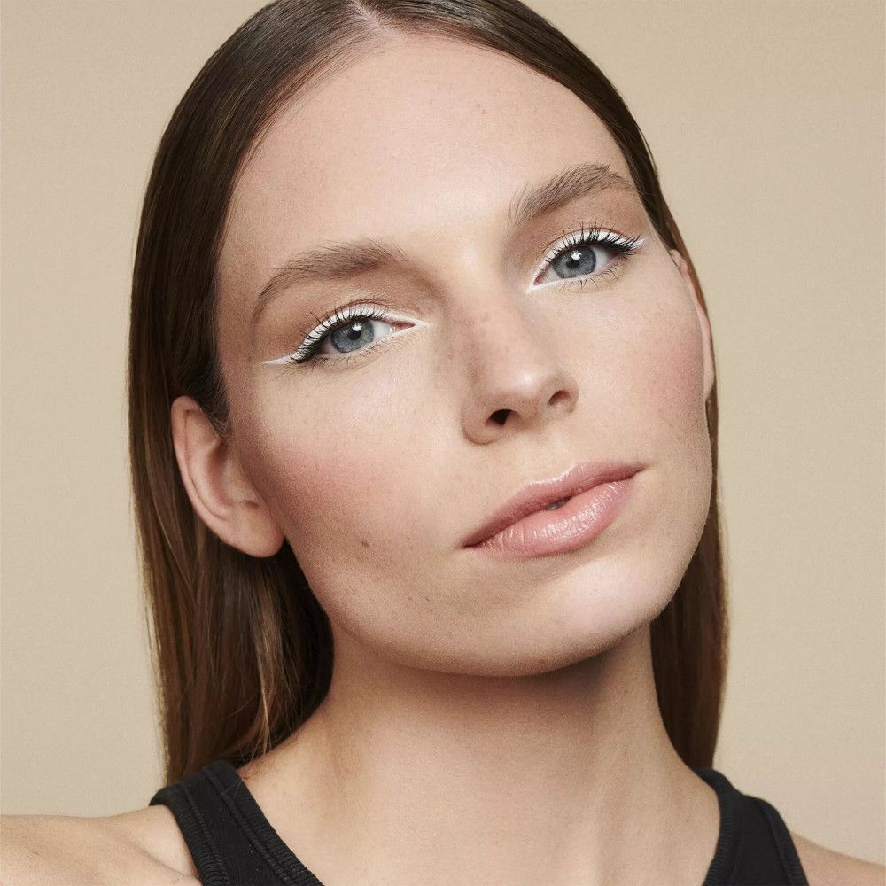 Nahaufnahme einer Frau mit hellem Teint, die weißen Eyeliner trägt. Ihre Augen sind blau und ihre Lippen sind leicht geschminkt. Der Hintergrund ist einfarbig.