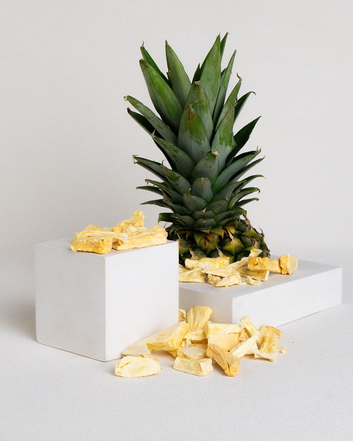 Frische Ananas mit grünen Blättern und gefriergetrockneten Ananasstücken auf weißen Würfeln. Einige Stücke liegen verstreut.