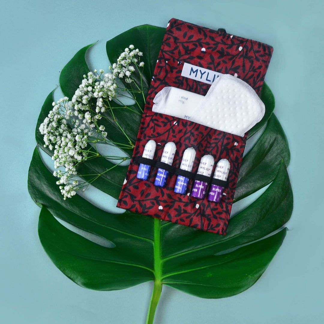 Offene, rot gemusterte Tasche mit Tampons und Binden. Auf einem grünen Blatt platziert. Tampons in verschiedenen Farben.