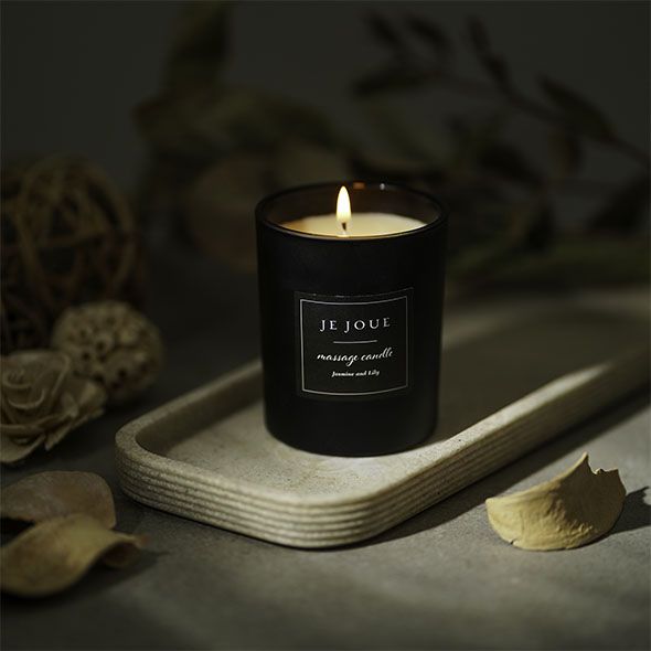 Brennende schwarze Kerze auf Tablett. Aufschrift: JE JOUE, massage candle, Jasmine and Lily.
