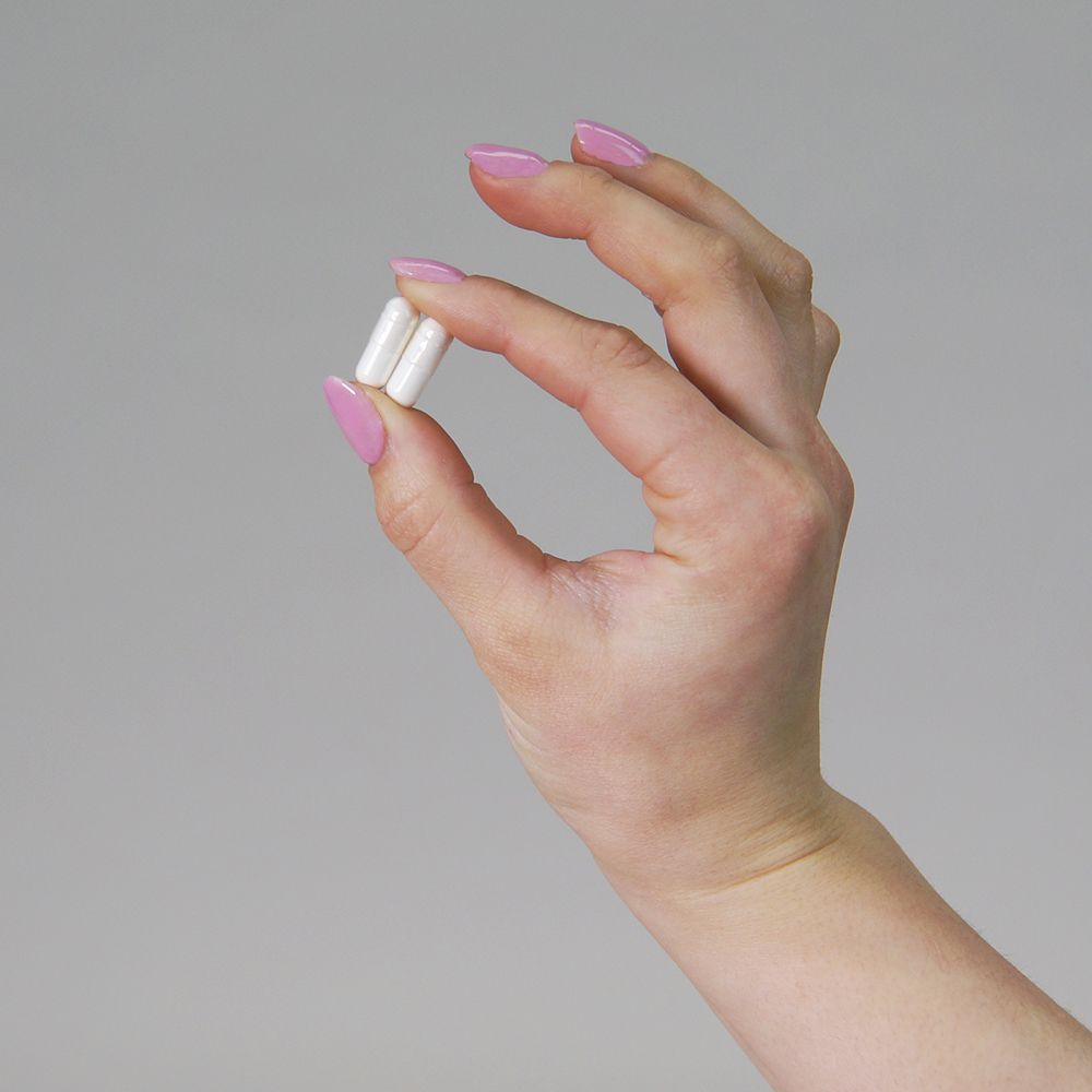 Hand hält zwei weiße Kapseln zwischen den Fingern. Rosa lackierte Fingernägel.