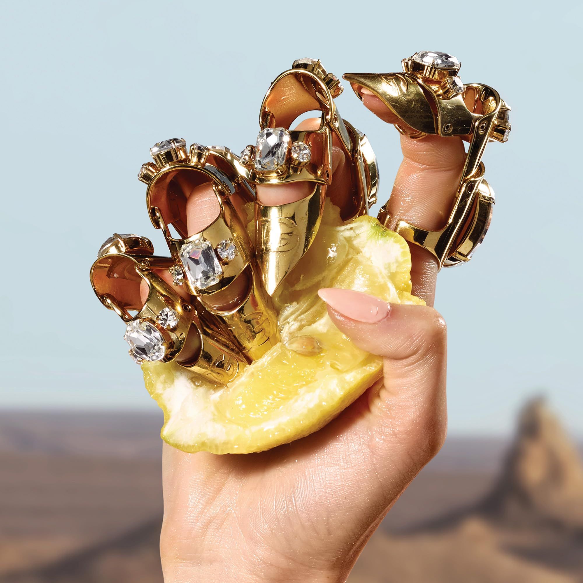 Hand mit goldfarbenen Ringen mit Schmucksteinen. Hand hält eine Zitronenhälfte. Hintergrund: Wüste und blauer Himmel.