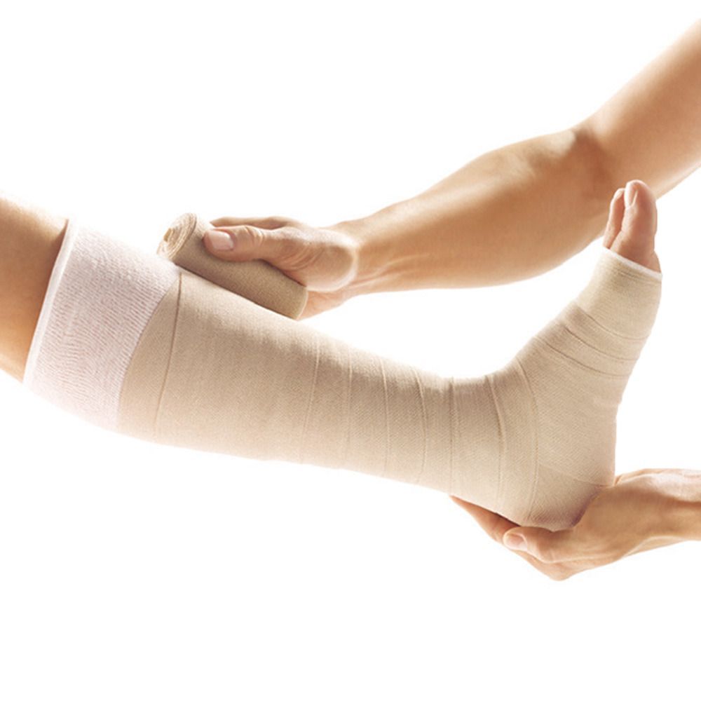 Bein mit Verband. Hände wickeln einen Verband um das Bein. Ein weiteres Verbandsmaterial liegt bereit.