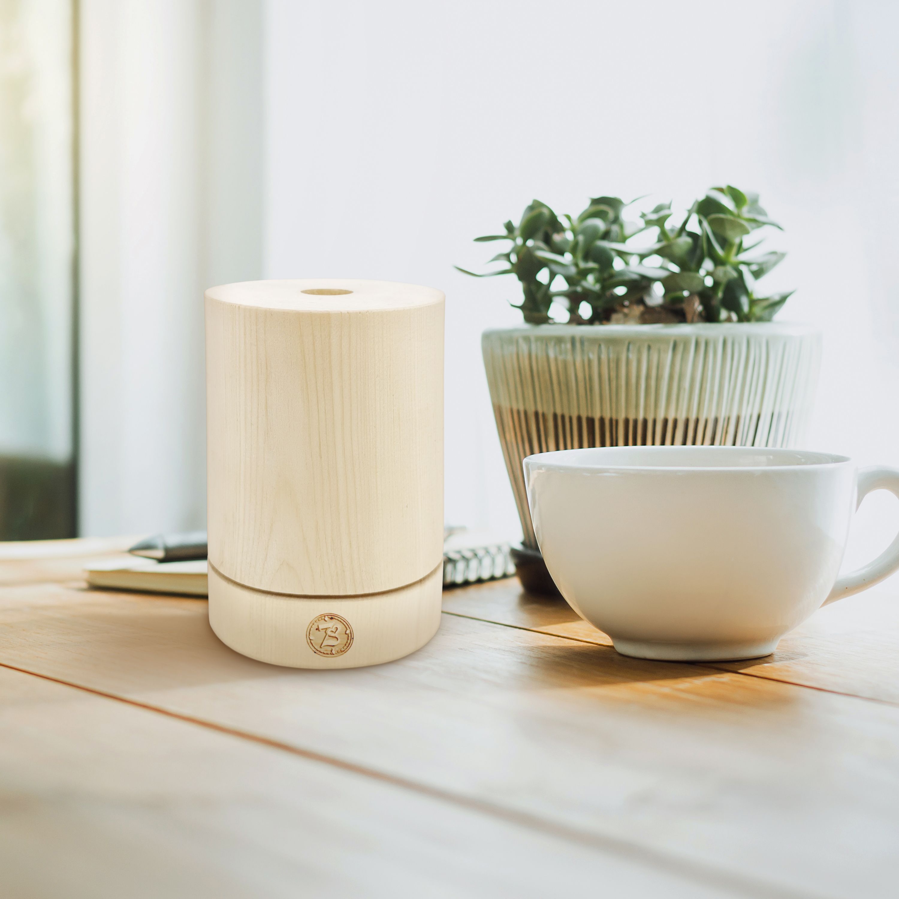 Ein Holzdiffusor steht auf einem Holztisch neben einer Kaffeetasse und einer Topfpflanze.