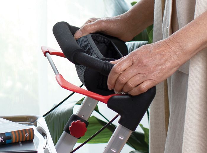 Person hält Handgriff eines Rollators. Roter Bremshebel, schwarzer Griff, grauer Rahmen. Teilansicht.