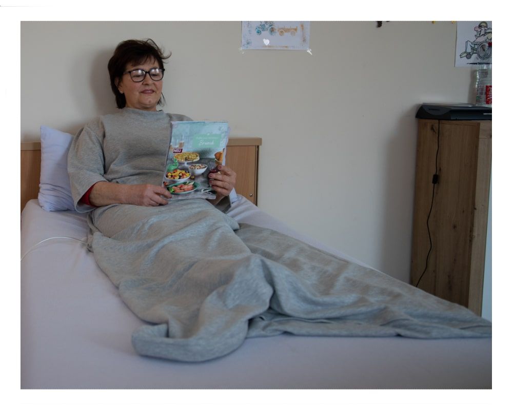Grauer Pflegeschlafsack, Frau sitzt im Bett und liest. Roter Saum am Ärmel. Buch mit farbigem Cover.