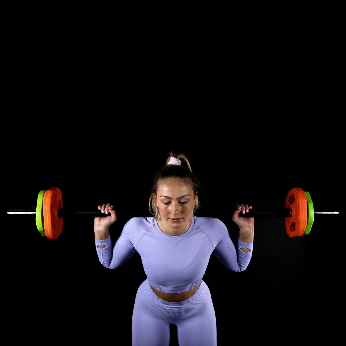 Frau in Sportkleidung mit Langhantel auf den Schultern. Orange und grüne Gewichtsscheiben.