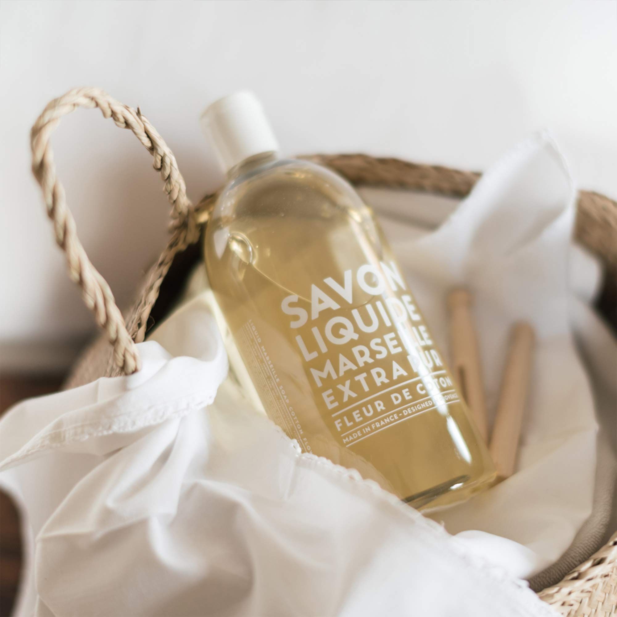 Transparente Flüssigseife in einer Flasche mit weißem Verschluss. Aufschrift: Savon Liquide, Marseille, Extra Pur, Fleur de Coton. Im Korb.