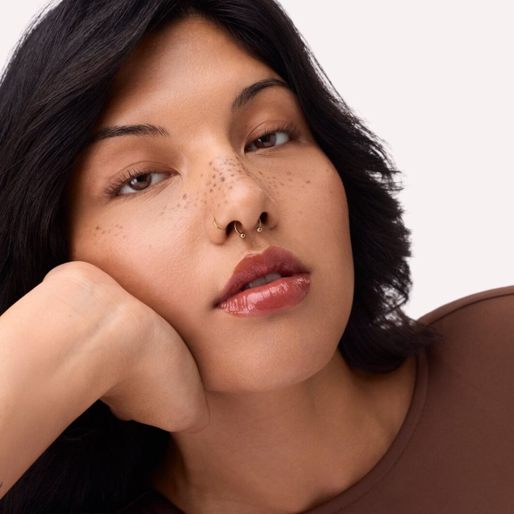 Porträt einer Frau mit Catrice Supreme Lippenfiller Gloss. Glänzende, orange-braune Lippen. Dunkles Haar, Sommersprossen, Nasenpiercing.