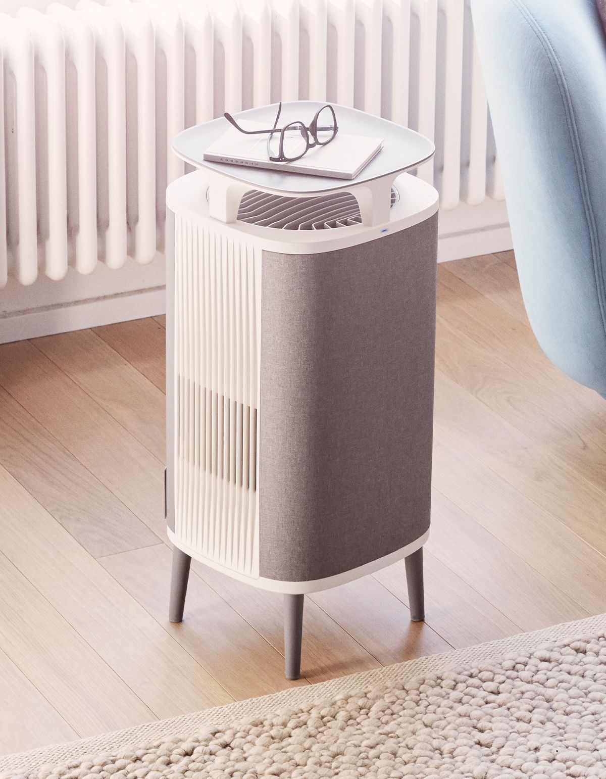 Luftreiniger in Wohnumgebung. Grauer Stoffbezug, weißer Rahmen, Beine. Brille und Notizen oben.