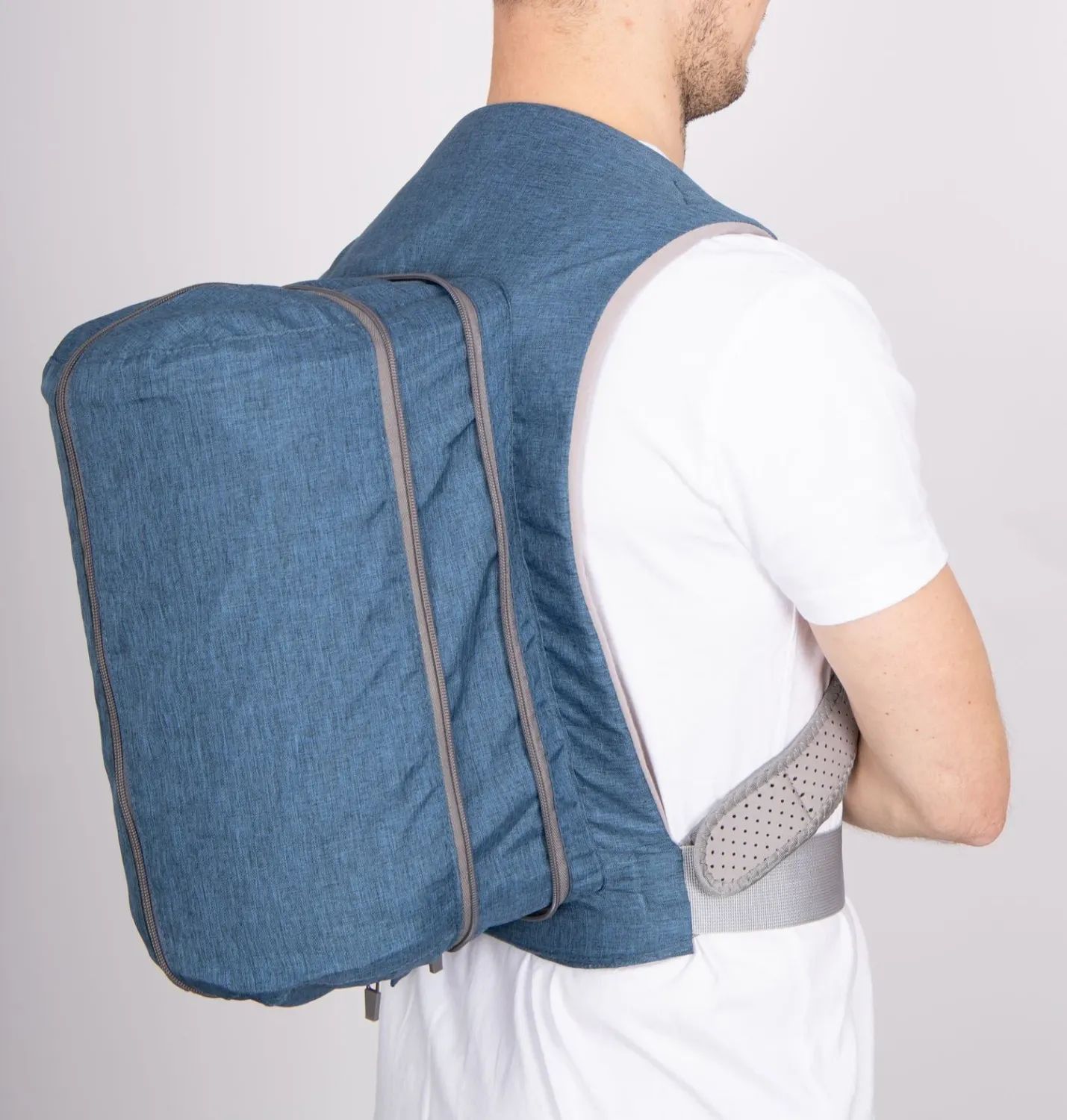 Blauer Rückenlageverhinderungsrucksack, getragen von einer Person. Graue Reißverschlüsse und Details. Person steht vor hellem Hintergrund.
