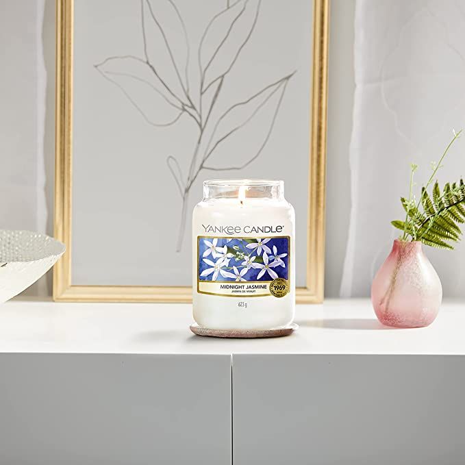 Duftkerze mit Etikett "Midnight Jasmine". Auf rosa Untersetzer. Im Hintergrund: Bild, Vase mit Zweig.