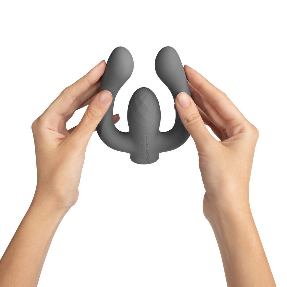 Grauer Vibrator, gehalten von zwei Händen. Das Produkt hat zwei gebogene Arme und einen geriffelten Mittelteil.
