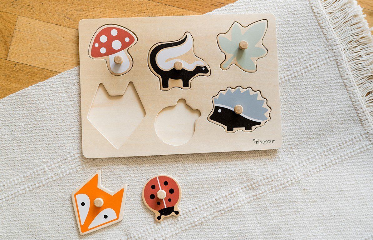 Holzpuzzle mit Waldtieren. Sechs farbige Puzzleteile: Pilz, Stinktier, Blatt, Fuchs, Marienkäfer, Igel. Zwei Puzzleteile daneben.