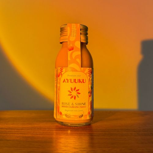 Gelbe Shot-Flasche auf Holz. Aufschrift: AYUUKU, RISE & SHINE, Ingwer, Kurkuma, Mango. Gelber Hintergrund mit Schatten.
