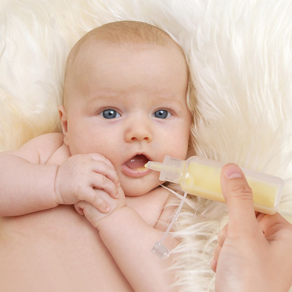 Baby erhält Flüssigkeit aus einem transparenten Behälter. Der Behälter wird von einer Hand gehalten.