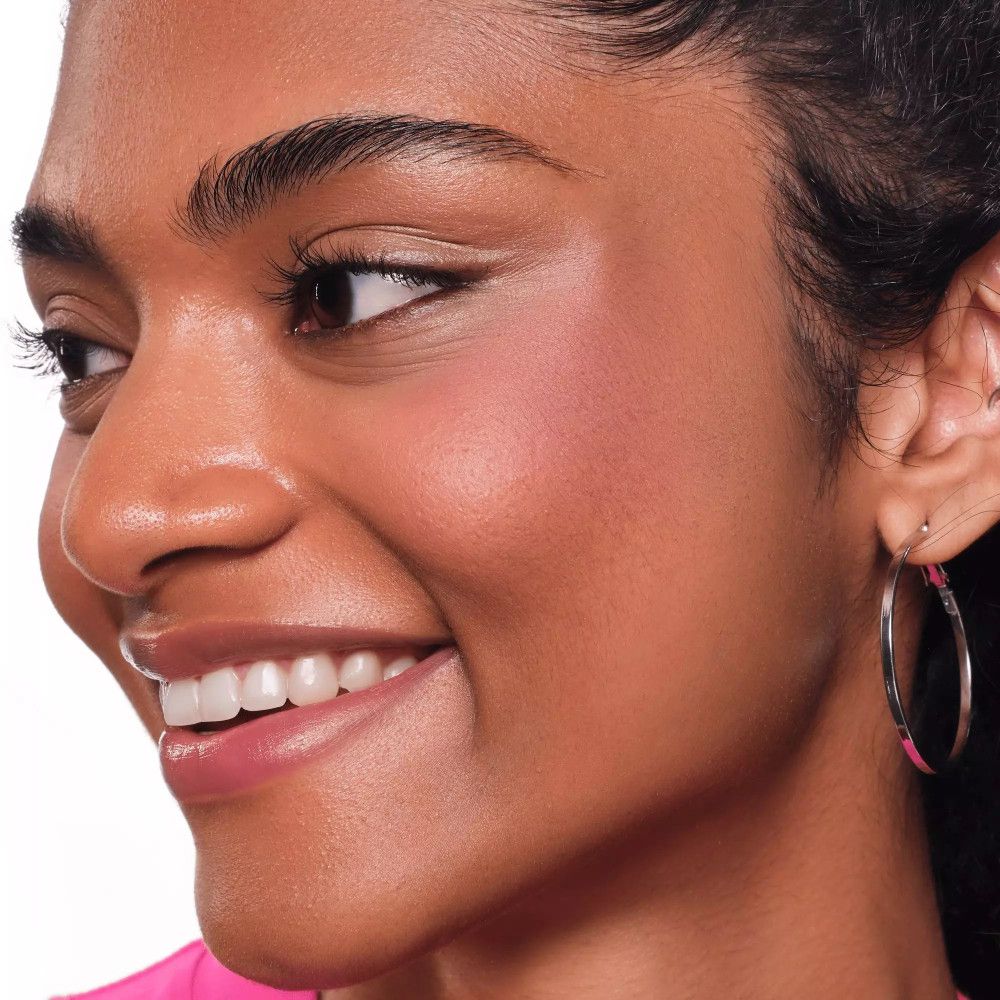 Nahaufnahme einer Frau mit rosa Rouge auf den Wangen. Sie trägt einen Ohrring und lächelt.