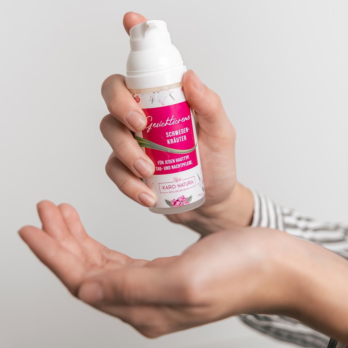 Hände halten Spender. Pinkfarbenes Etikett. Text: Gesichtscreme, Schwedenkräuter, Karo Natura.