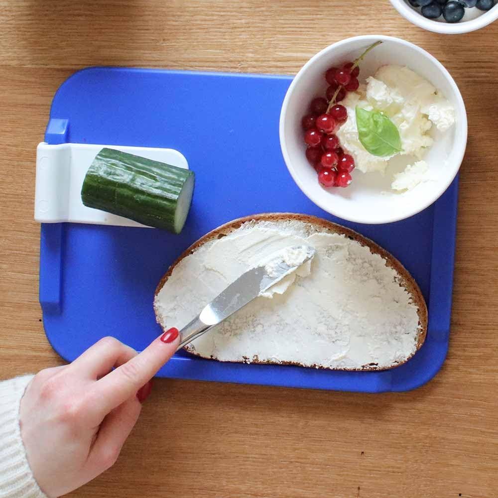 Blaues Schneidebrett mit Gurkenscheibe, Brot und Butter. Eine Hand bestreicht das Brot.