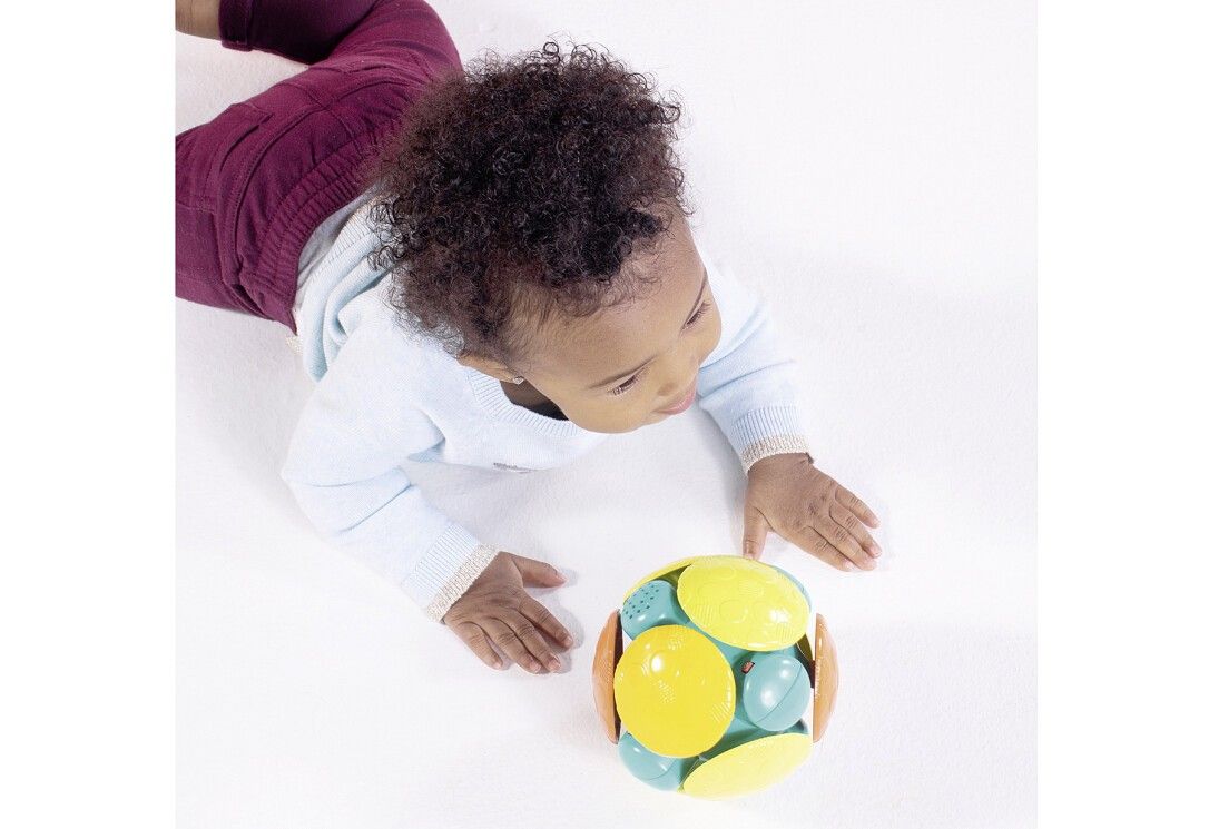 Baby betrachtet einen bunten Ball. Der Ball hat gelbe, blaue, orange und grüne Elemente. Das Baby liegt auf dem Bauch.