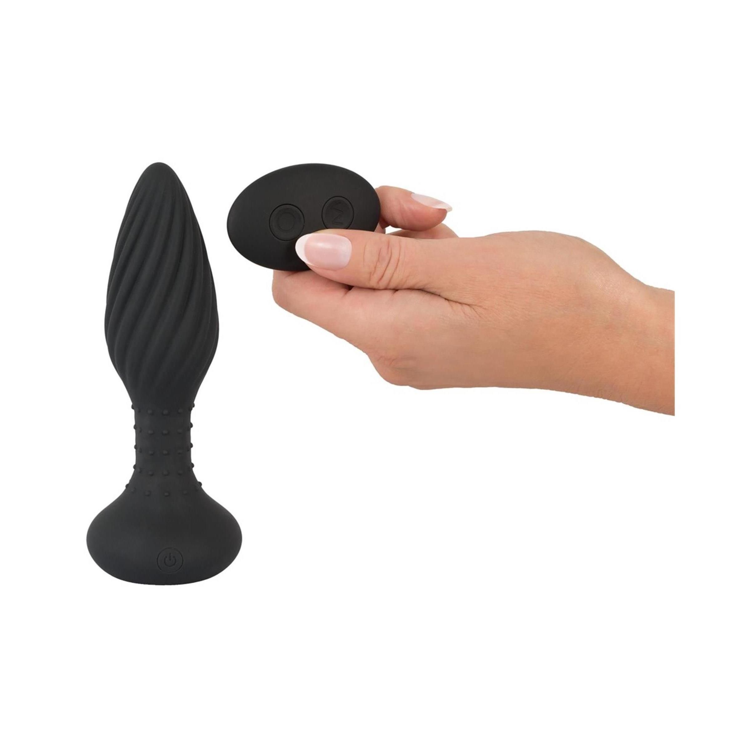 Eine Hand hält eine Fernbedienung. Daneben ein schwarzer Anos RC Butt Plug. Der Hintergrund ist weiß.