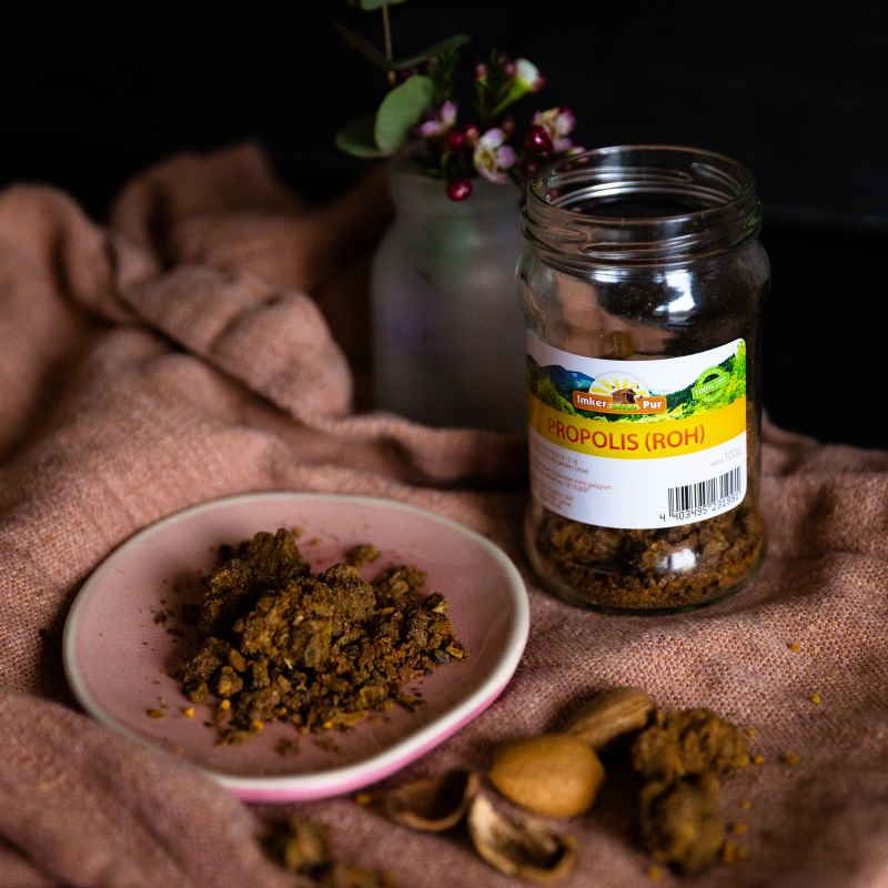 Propolis auf rosa Teller, neben Glasbehälter. Glasbehälter mit Etikett. Mandeln und Holzschaufel daneben. Textiluntergrund.