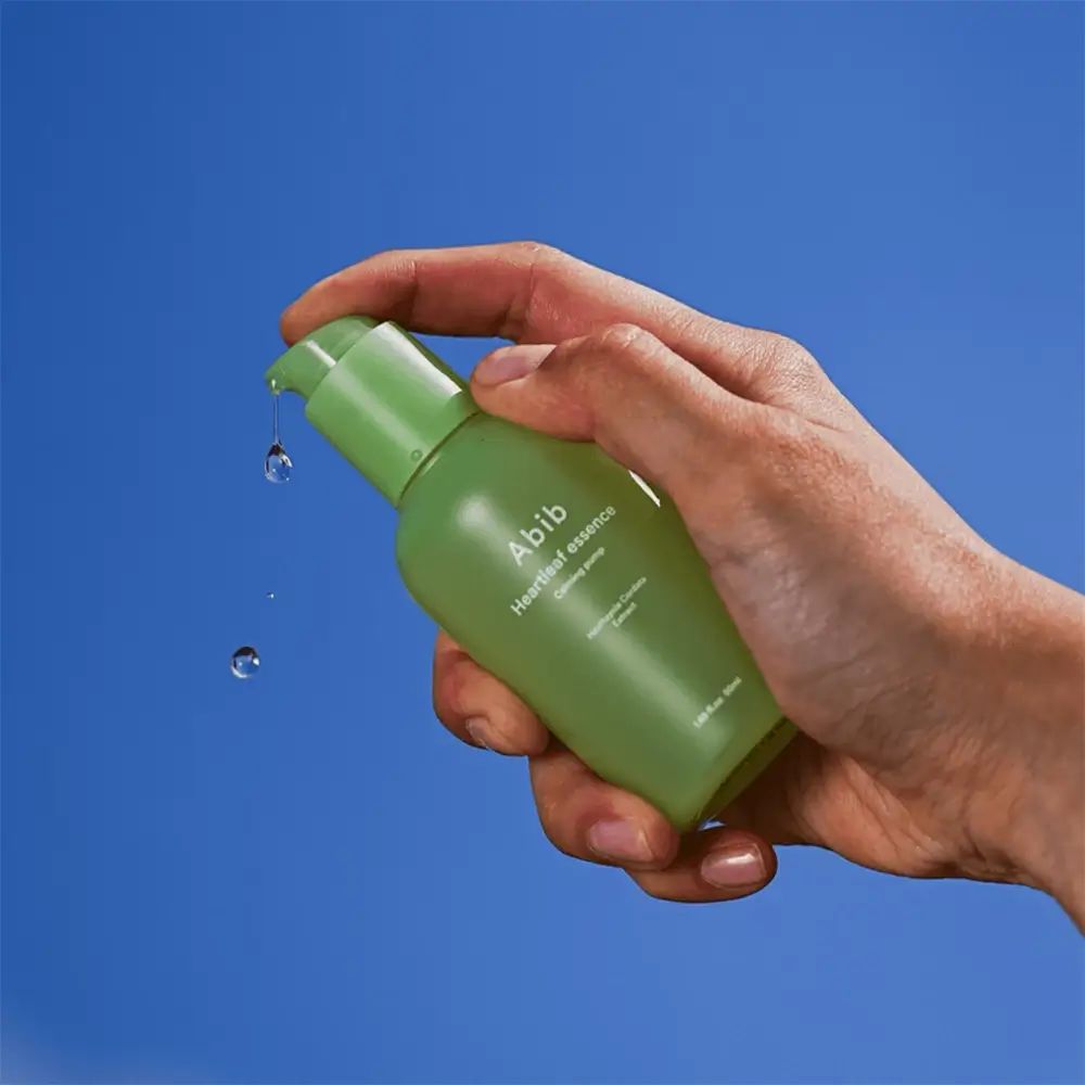 Grüne Flasche mit Pumpverschluss in einer Hand. Tropfen fallen. Aufschrift: Abib Heartleaf essence Calming pump.