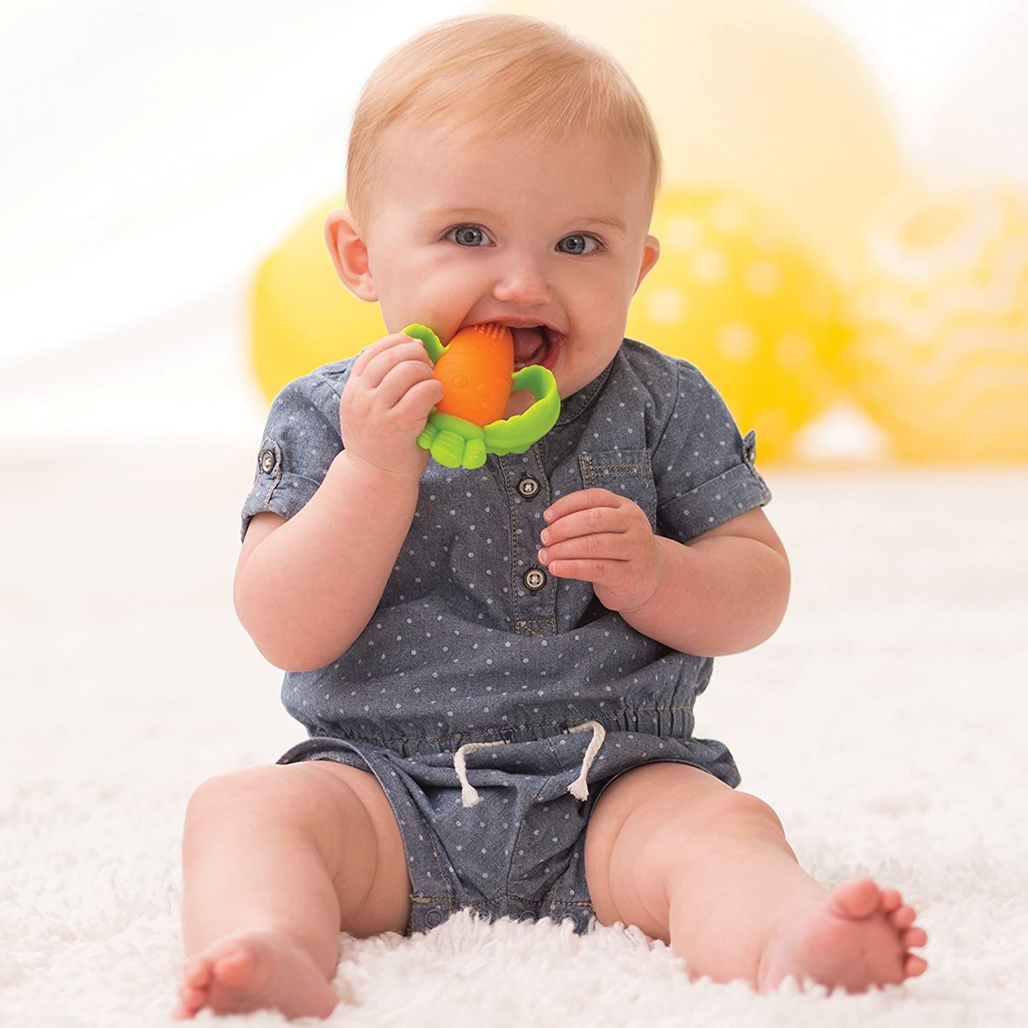 Baby mit blauem Overall, der einen orangefarbenen und grünen Karotten-Beißring hält und in den Mund nimmt. Auf weißem Teppich.