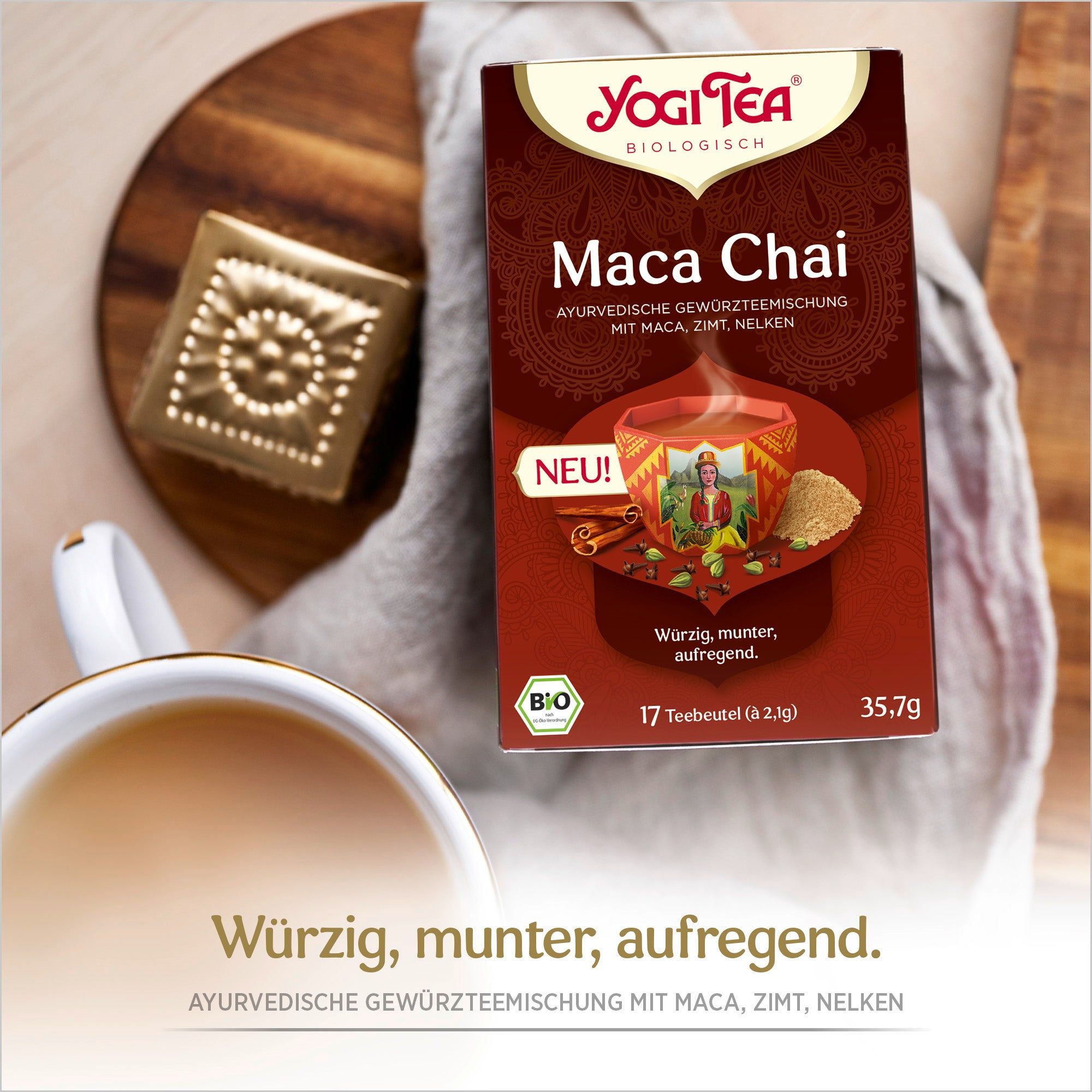 Yogi Tea Maca Chai auf Holzbrett. Braune Verpackung, Tasse Tee, Gebäck. Text: Würzig, munter, aufregend.