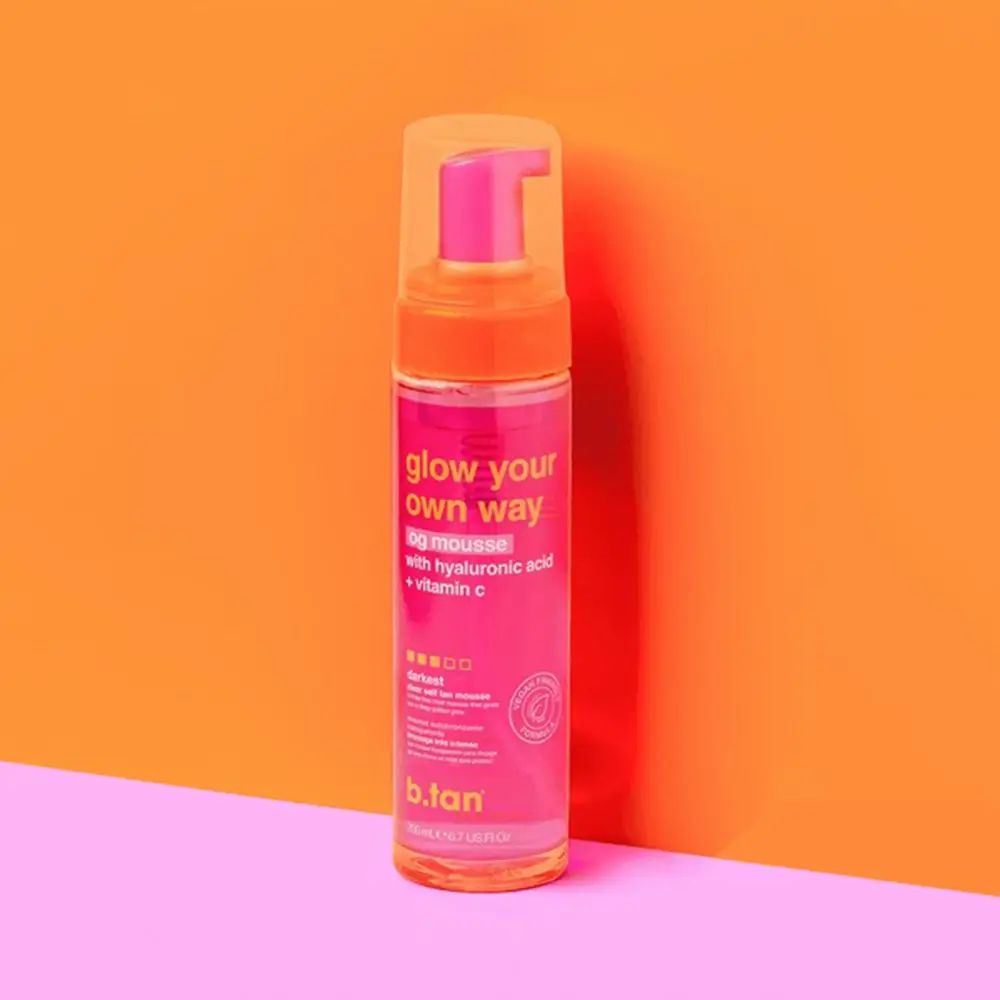 Rosa Flasche mit orangefarbenem Deckel. Aufschrift: glow your own way, og mousse, mit Hyaluronsäure + Vitamin C. Marke: b.tan.