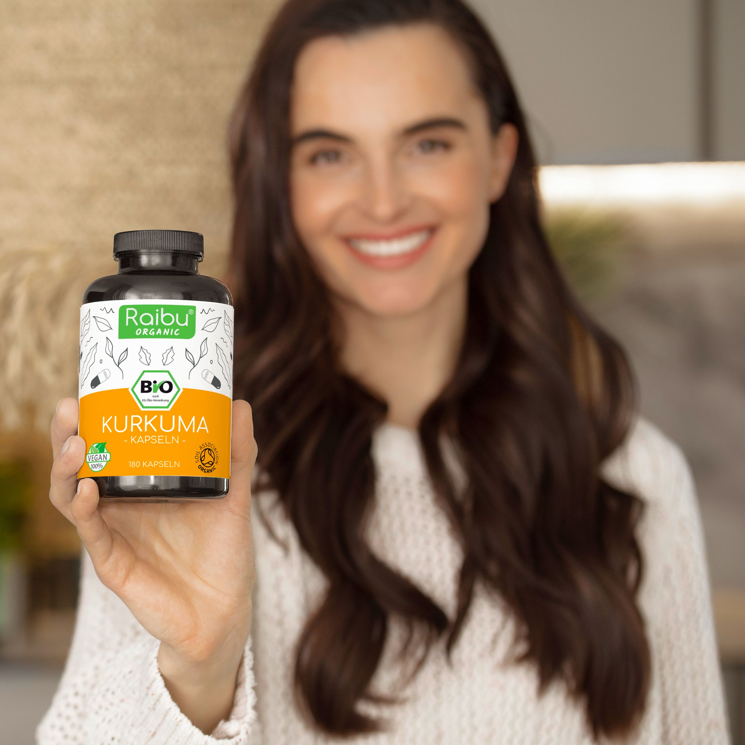 Frau hält eine Flasche. Aufschrift: Raibu Organic, Bio, Kurkuma, 180 Kapseln. Siegel: Vegan, Oil Association Organic. Frau lächelt.