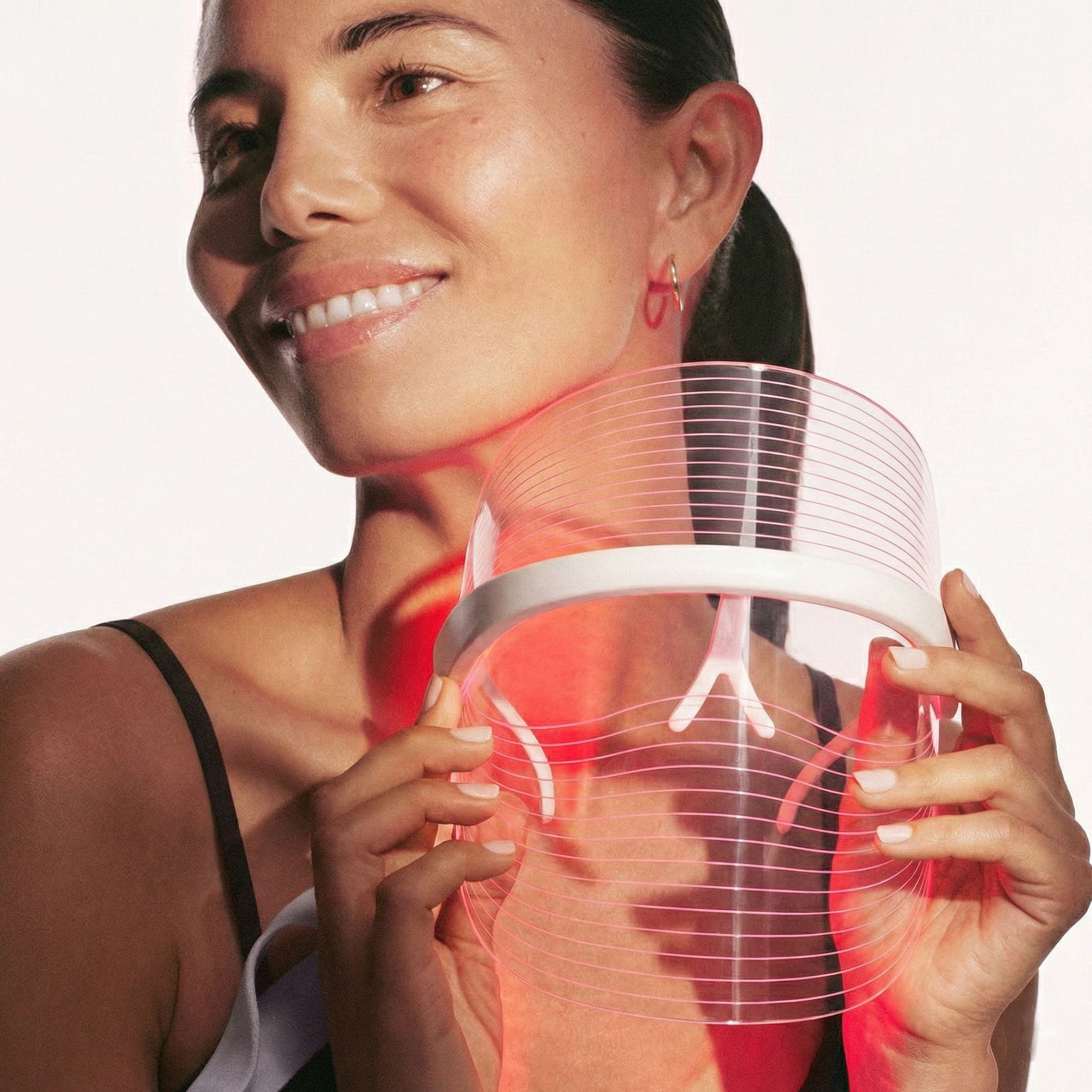 Frau hält eine weiße und transparente LED-Gesichtsmaske mit roten Lichtern, die auf ihren Halsbereich gerichtet sind.