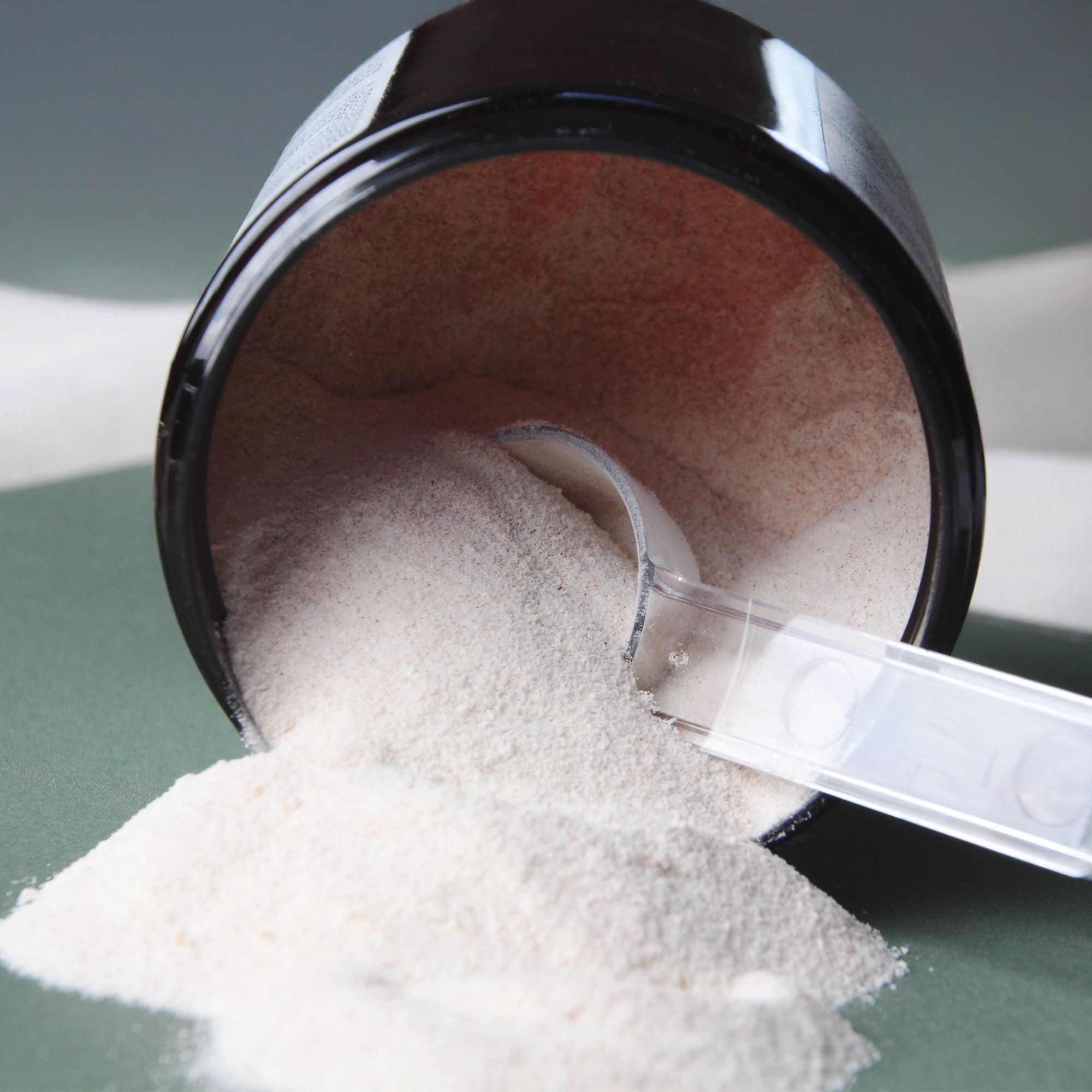 Geöffnete Dose mit Pulver und Messlöffel. Pulver liegt daneben. Dose und Löffel sind weiß.