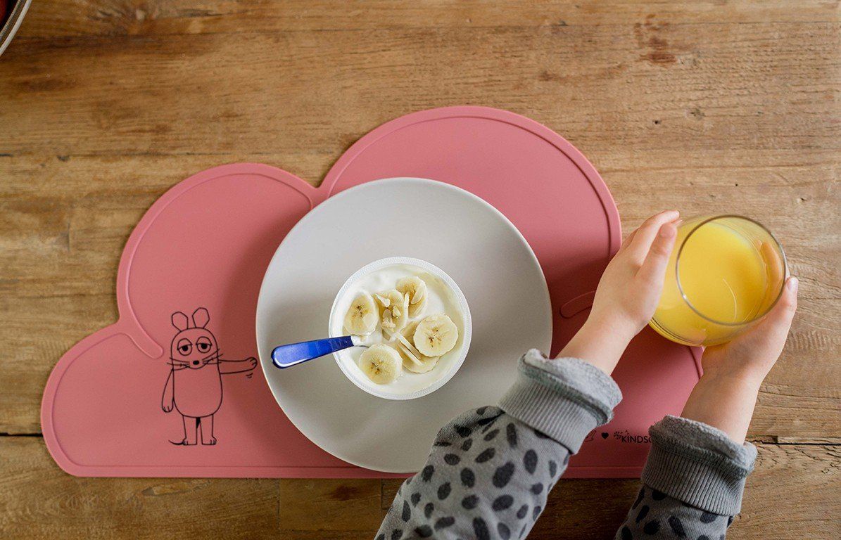 Rosa Platzdeckchen in Wolkenform mit Essen. Teller mit Joghurt und Banane, Glas Saft.