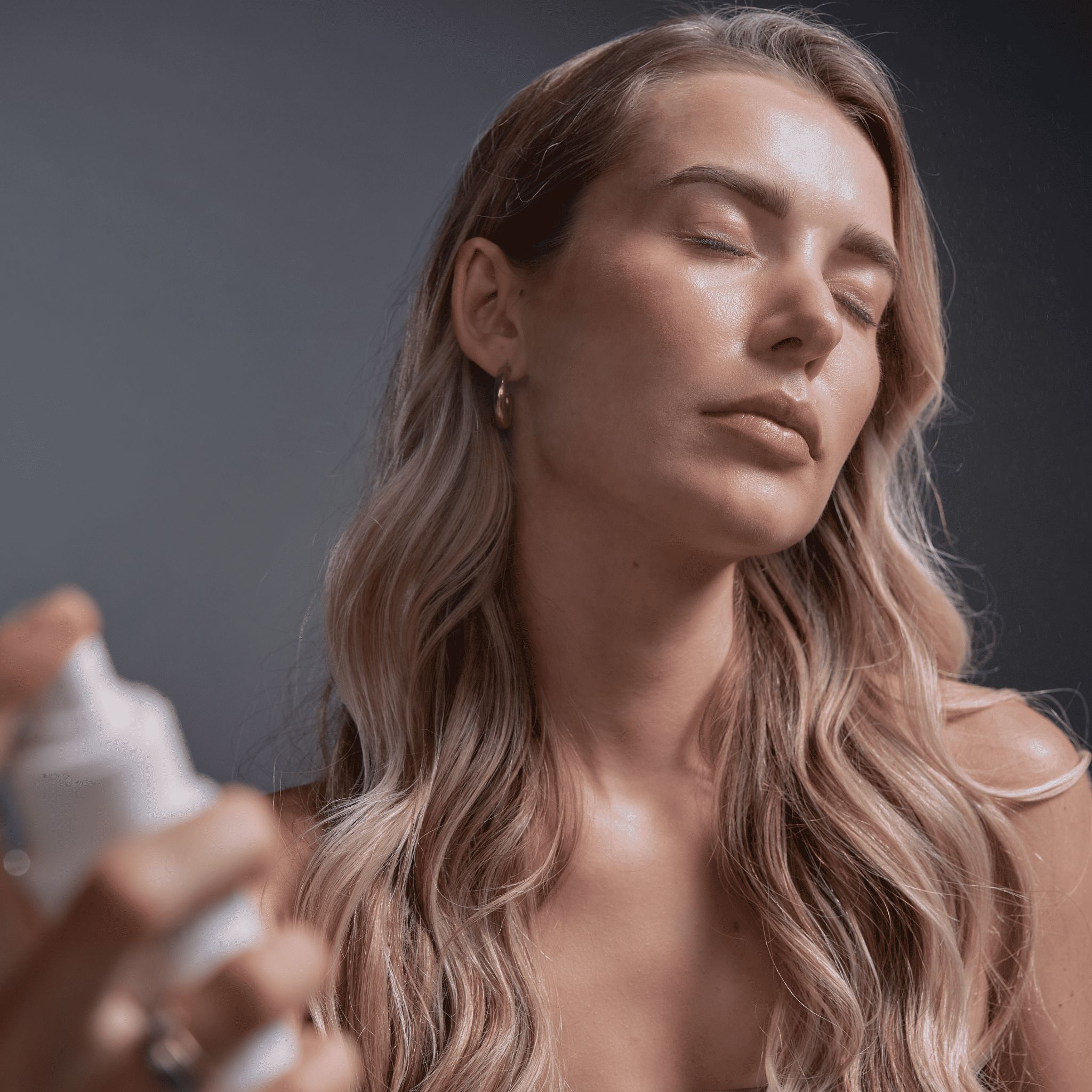 Frau sprüht Gesichtsspray auf ihr Gesicht. Blondes Haar, geschlossene Augen. Sprayflasche in der Hand.