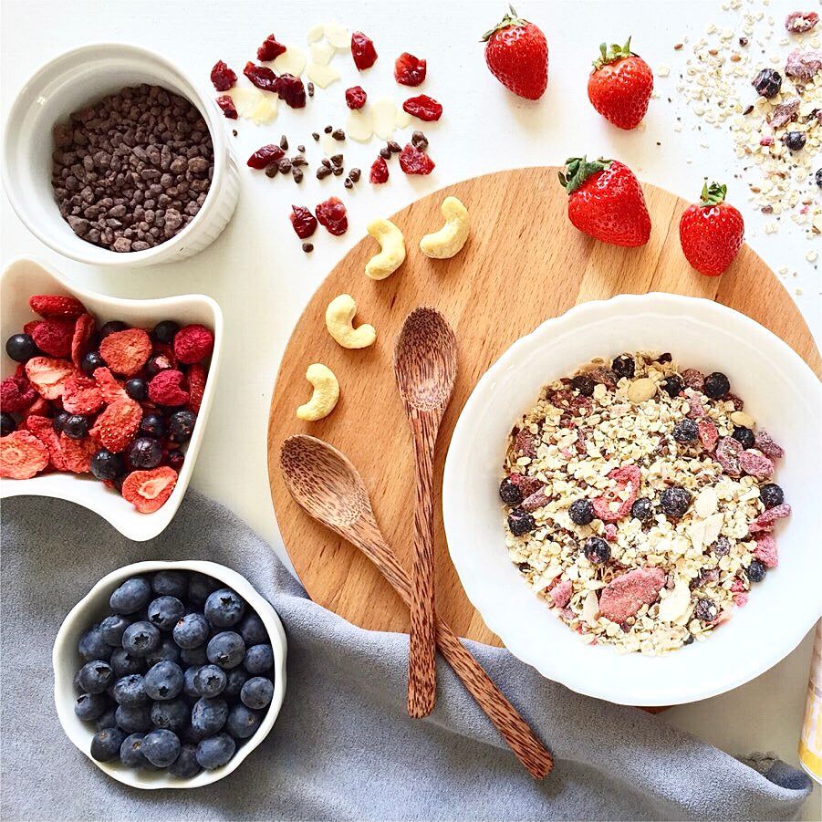 Verschiedene Zutaten auf Holzbrett: Blaubeeren, Erdbeeren, Cashewnüsse, Schokostückchen, Himbeeren. Zwei Holzlöffel.