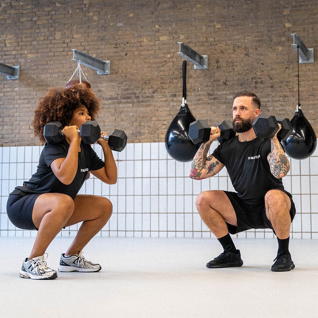 Zwei Personen machen Kniebeugen mit Kurzhanteln. Sie tragen schwarze Shirts mit dem Logo "MATCHU".
