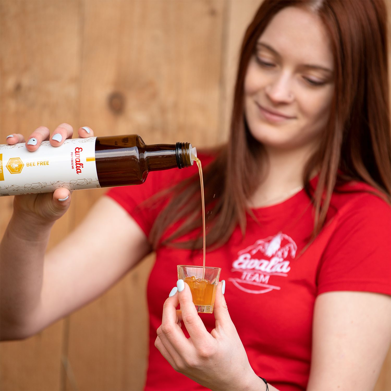 Frau gießt Flüssigkeit aus einer Flasche in ein Glas. Flasche mit Etikett: Ewalia, BEE FREE. Frau trägt rotes T-Shirt.
