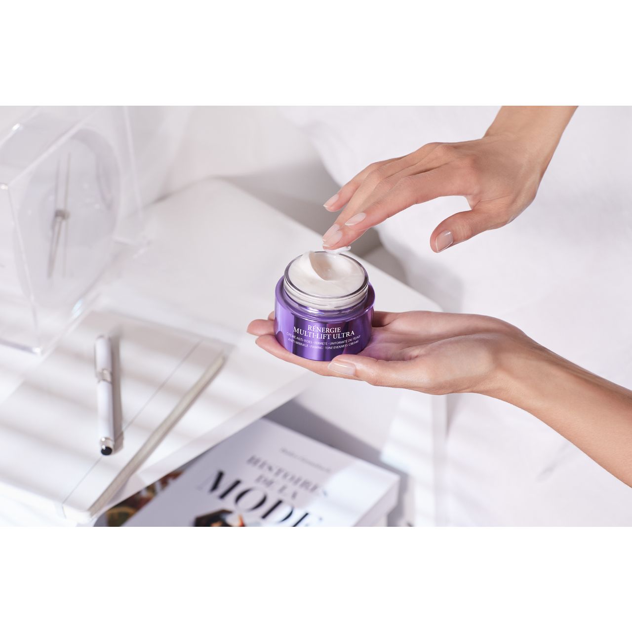 Hände halten einen violetten Tiegel mit Creme. Aufschrift: Rénergie Multi-Lift Ultra. Im Hintergrund: Buch, Uhr.