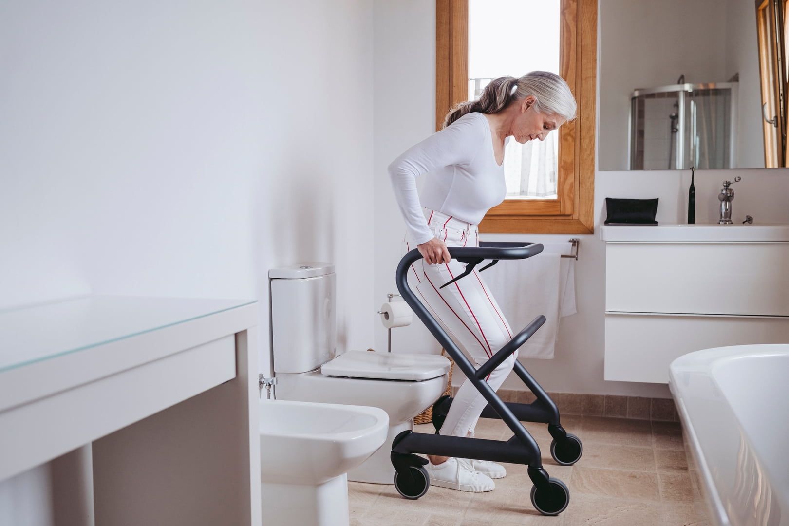 Frau im Badezimmer, stützt sich auf Rollator. Rollator mit vier Rädern, schwarzer Rahmen.