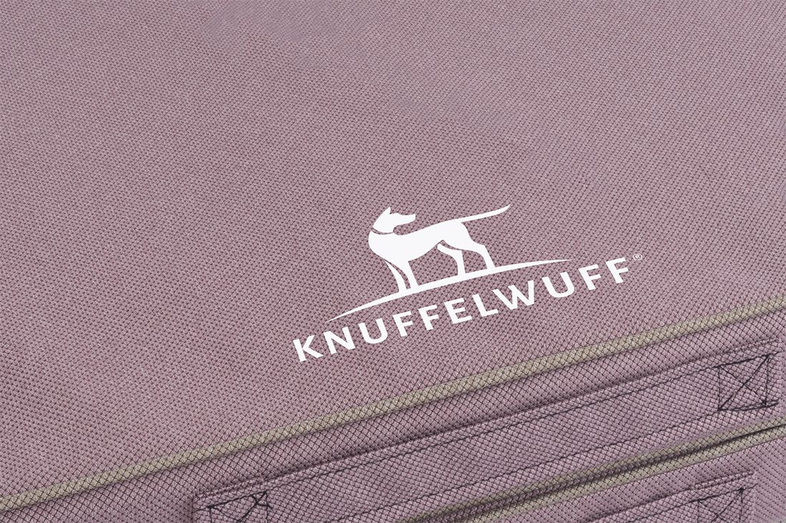 Detailansicht des Knuffelwuff-Logos auf fliederfarbenem Stoff. Logo: weißer Hund auf Hügel, Schriftzug.