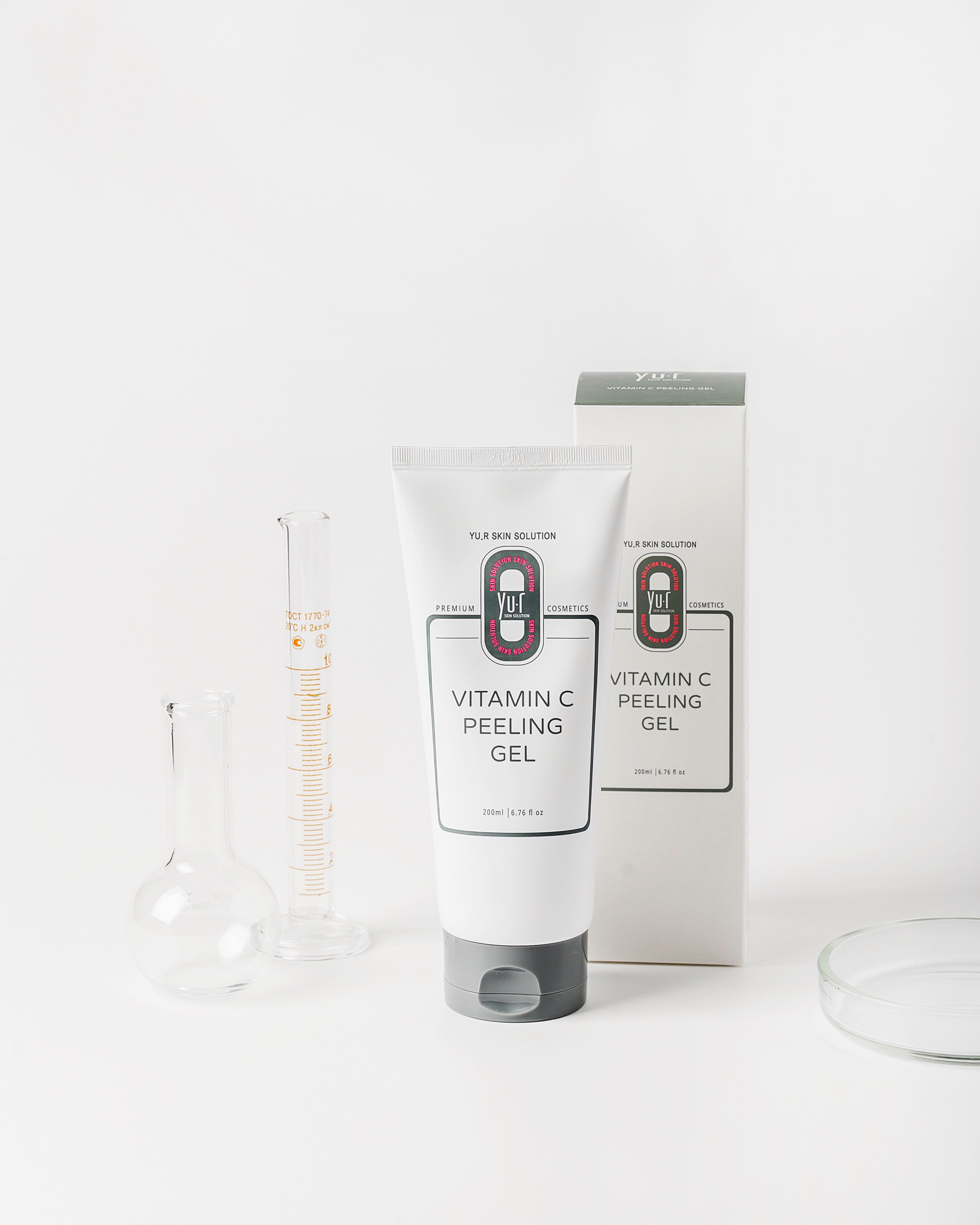 Weiße Tube mit grauer Kappe und Aufschrift Vitamin C Peeling Gel. Daneben Verpackung und Laborutensilien.
