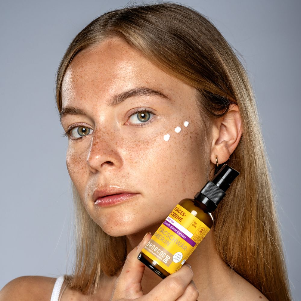 Frau mit benecosBIO Tagescreme BIO-Olive Flakon in der Hand, weiße Creme-Punkte auf der Wange.