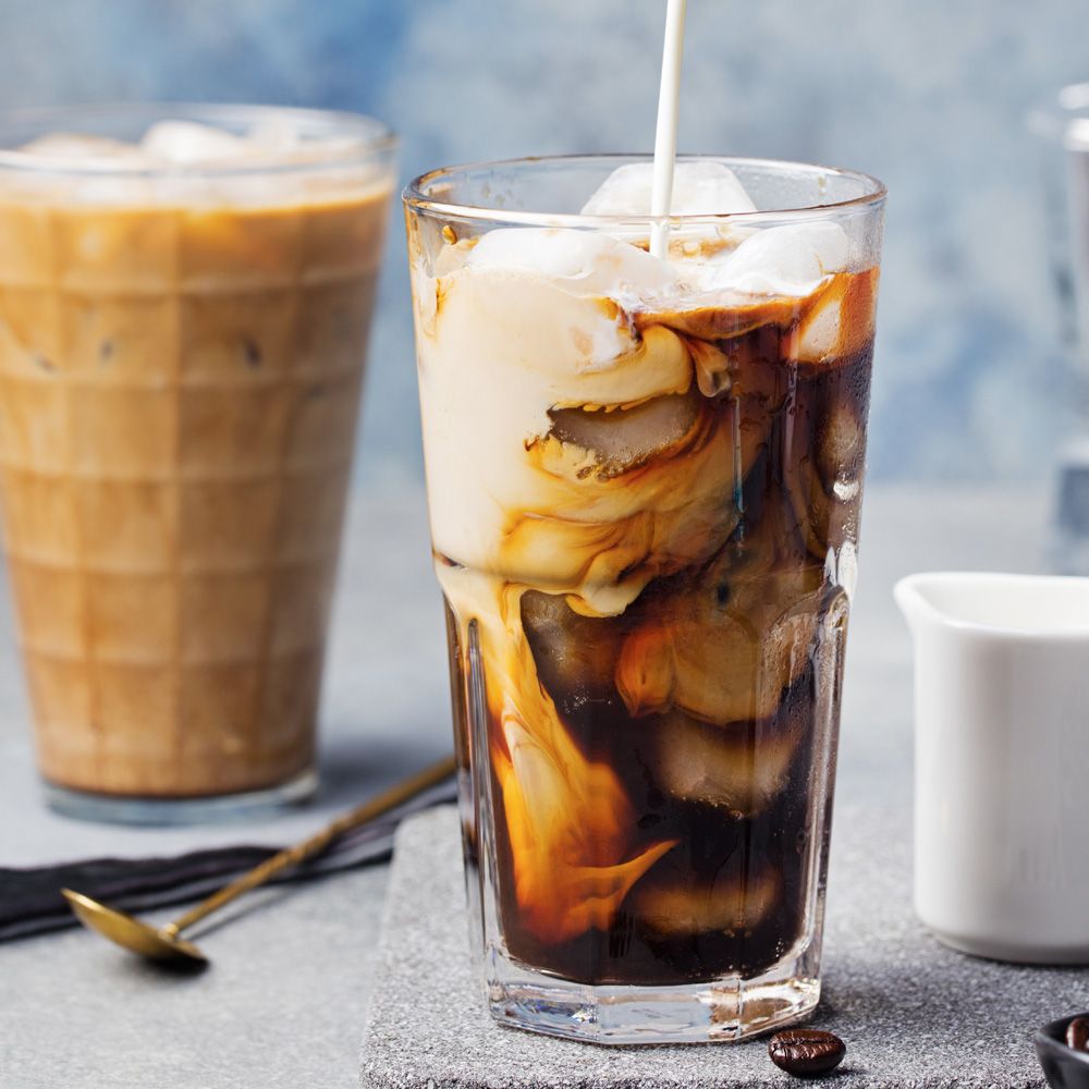 Glas mit Getränk, Eiswürfeln und Milch. Kaffeebohnen und Milchkännchen im Hintergrund. Flüssigkeiten mischen sich im Glas.