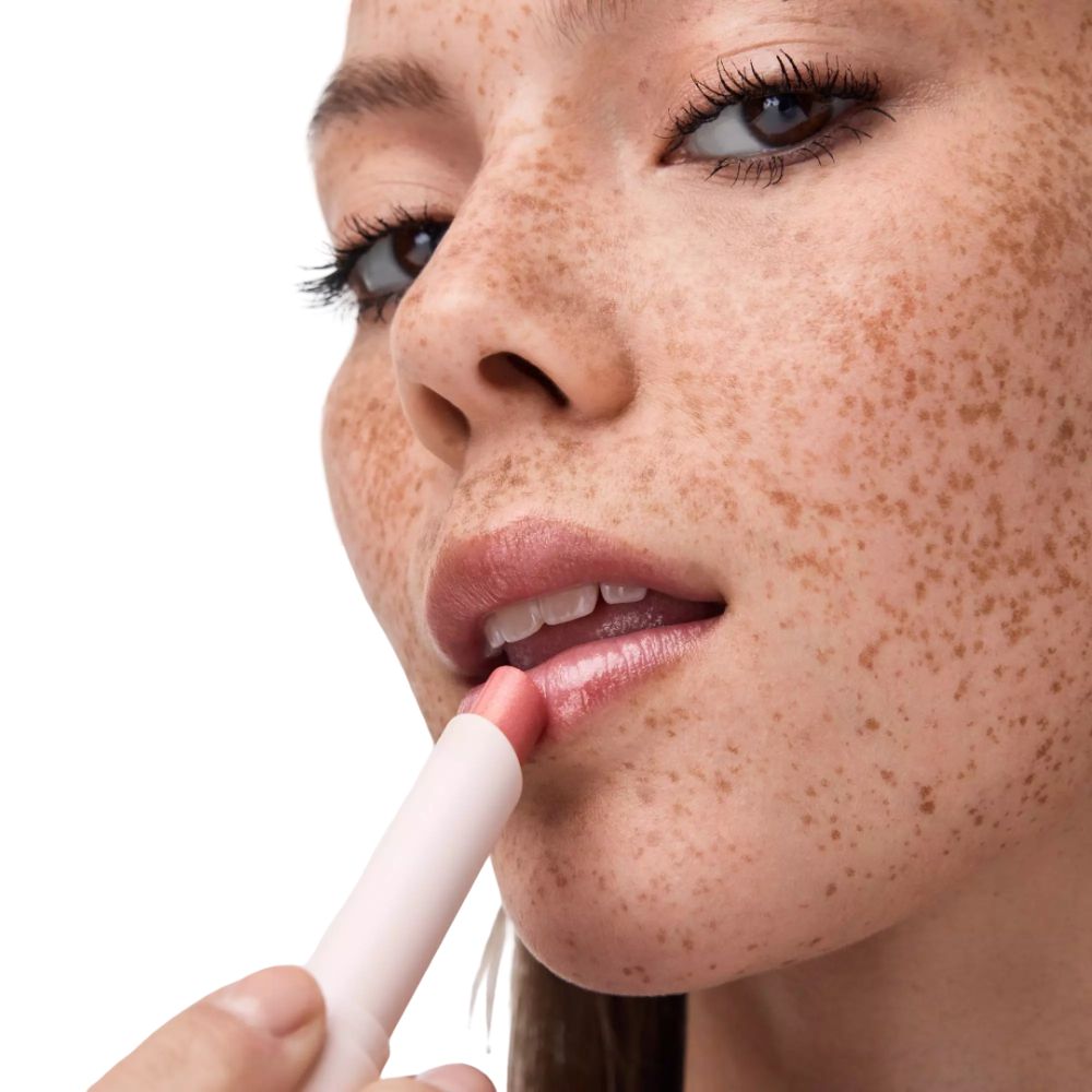 Nahaufnahme einer Frau mit Sommersprossen, die einen hellrosa Lippenbalsam aufträgt.